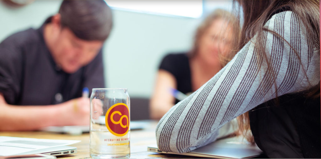 Three people are seated at a table in a meeting setting, partially visible, focusing on their work. A glass with a yellow and red logo is in the foreground, while notebooks and laptops are present. The scene appears collaborative and professional.