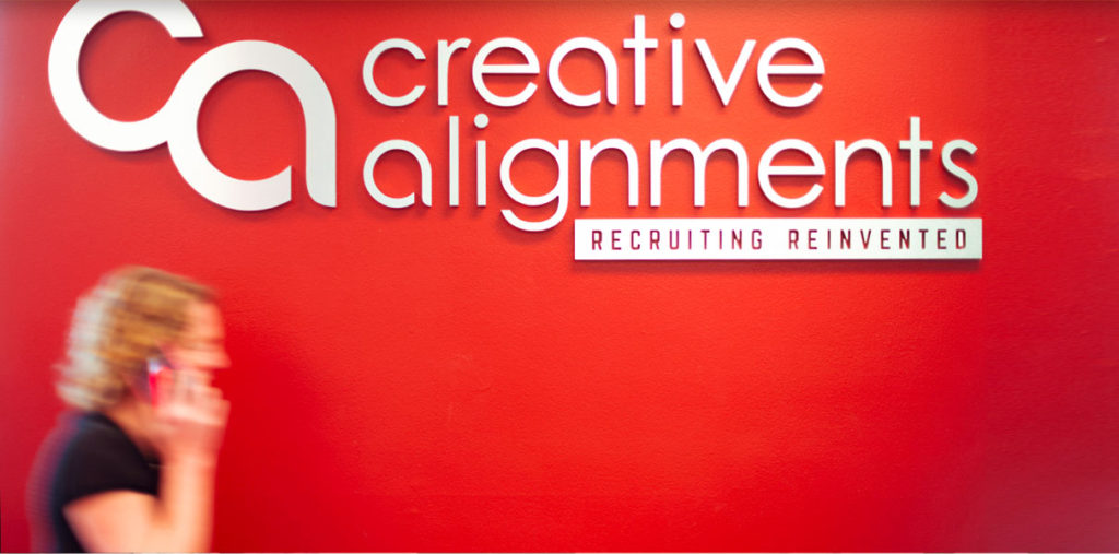 A person with curly hair is walking and talking on a phone in front of a red wall that has the text "Creative Alignments" and "Recruiting Reinvented" displayed in large letters.