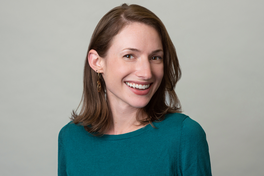 A woman with shoulder-length brown hair, wearing a teal top and gold earrings, smiles at the camera against a plain light gray background.