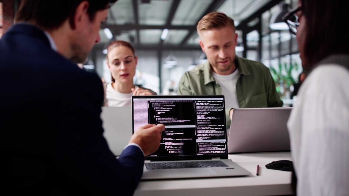 Four people sit at a table in a modern office, focused on laptops. One laptop screen shows lines of code, while a man in a dark suit gestures toward it, discussing with the group.