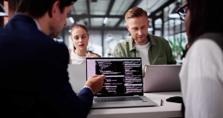 Four people sit at a table in a modern office, focused on laptops. One laptop screen shows lines of code, while a man in a dark suit gestures toward it, discussing with the group.