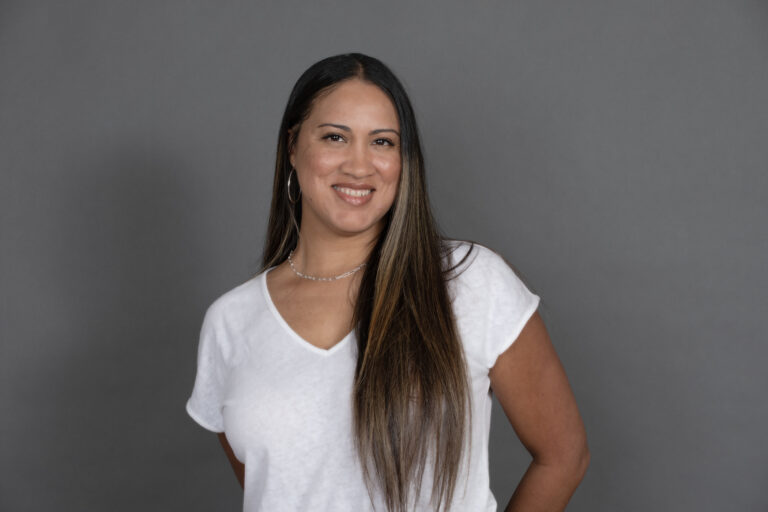 A woman with long straight hair, wearing a white V-neck shirt and hoop earrings, smiles while standing against a plain gray background.