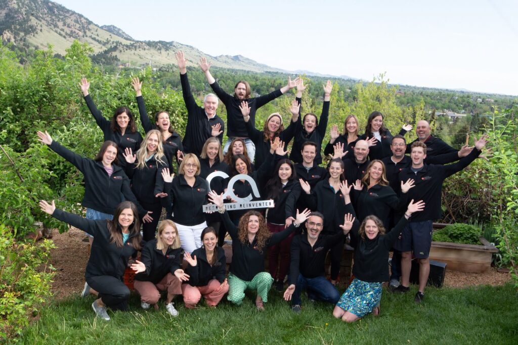 A group of people outdoors in front of green bushes and mountains, all wearing black jackets and raising their arms, smiling cheerfully, with some holding a sign that reads "Relivcenter.
