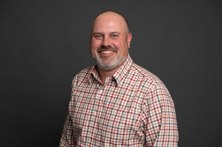 A smiling man with a beard and shaved head wearing a red, white, and blue plaid shirt stands against a plain dark gray background.
