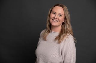 A woman with long, blonde hair smiles at the camera. She is wearing a light-colored sweater and hoop earrings, standing against a plain dark background.
