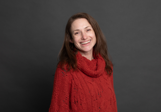 A woman with long brown hair, wearing a textured red sweater, smiles warmly while standing in front of a plain dark gray background.
