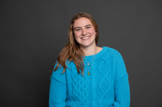 A young woman with long, light brown hair smiles at the camera. She is wearing a bright blue, cable-knit sweater and gold necklaces, with a dark gray background behind her.