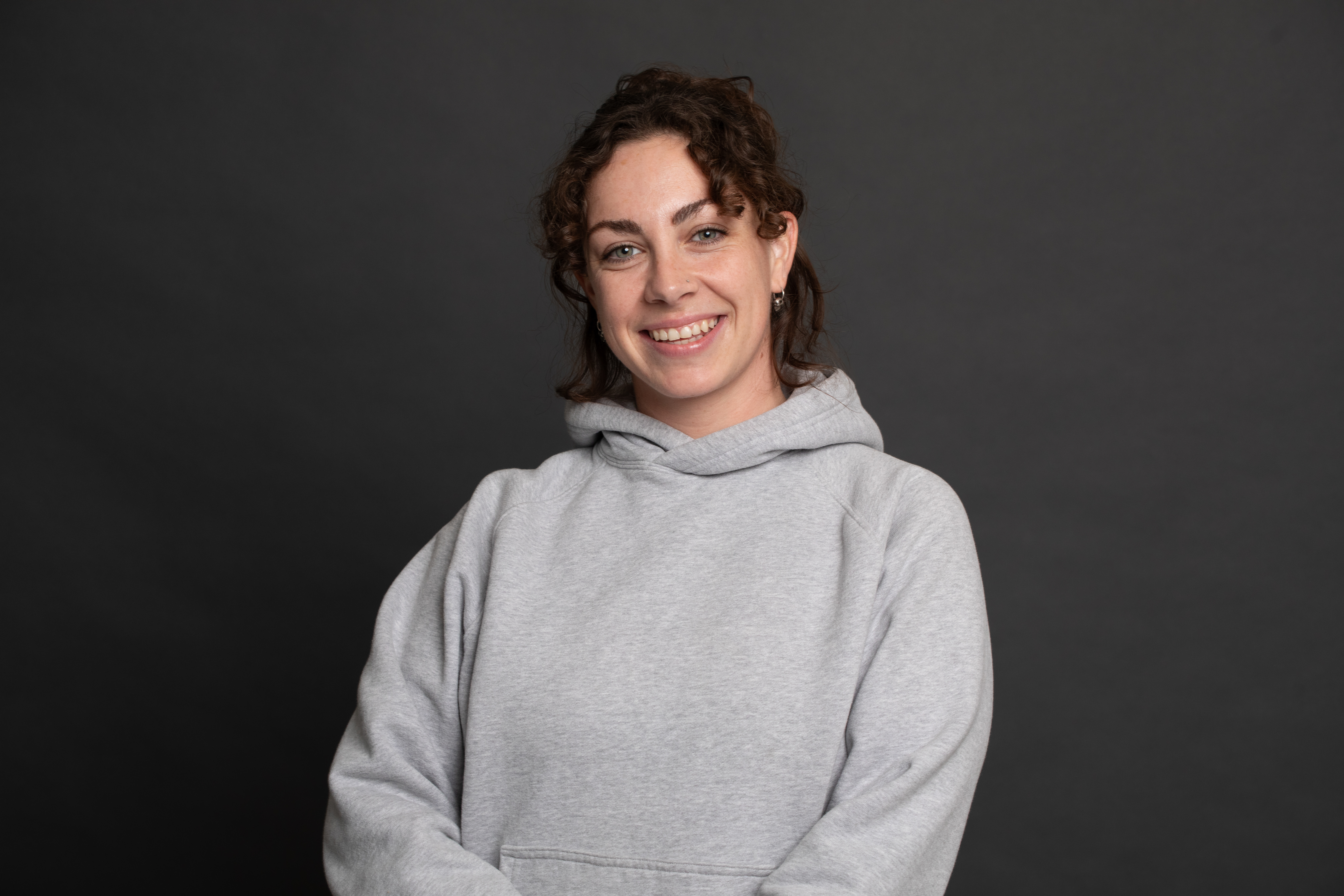 A woman with curly brown hair wearing a light gray hoodie smiles while standing against a plain dark gray background.