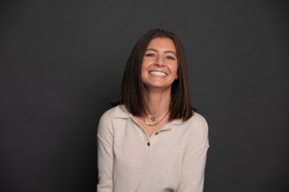 A woman with straight brown hair and a light beige sweater smiles while sitting in front of a plain dark background.