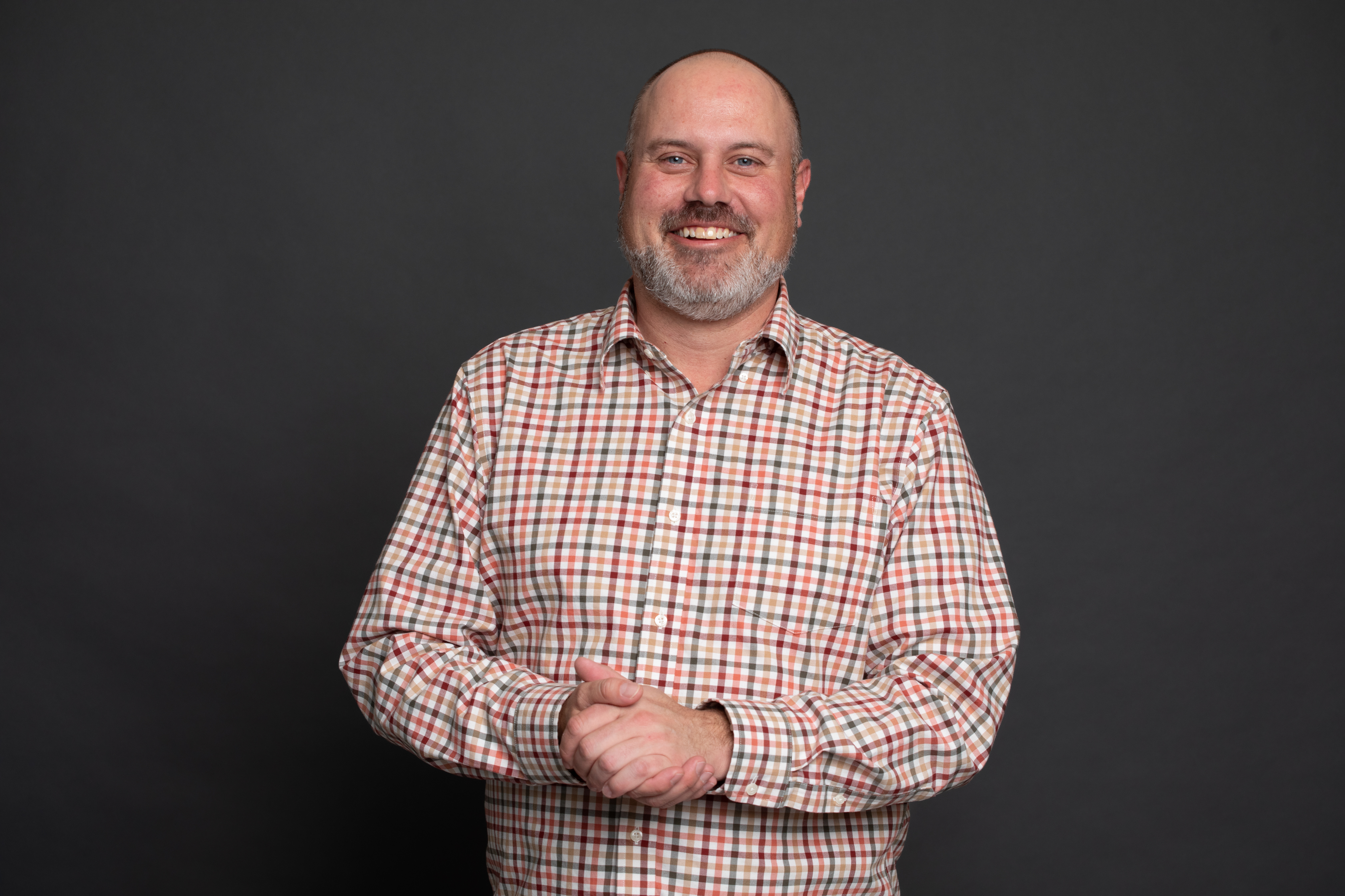 A man with a bald head and gray beard smiles, standing against a plain dark background. He is wearing a red, white, and beige plaid button-down shirt and has his hands clasped in front of him.