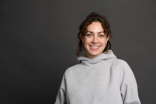 A young woman with curly brown hair smiles at the camera. She is wearing a light gray hoodie and stands in front of a plain dark gray background.