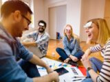 Four people sit on the floor laughing, each with a sticky note on their forehead. Papers, a tablet, and charts are spread in front of them, suggesting a playful group activity or team-building exercise in a bright room.