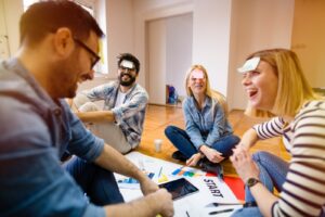 Four people sit on the floor laughing, each with a sticky note on their forehead. Papers, a tablet, and charts are spread in front of them, suggesting a playful group activity or team-building exercise in a bright room.