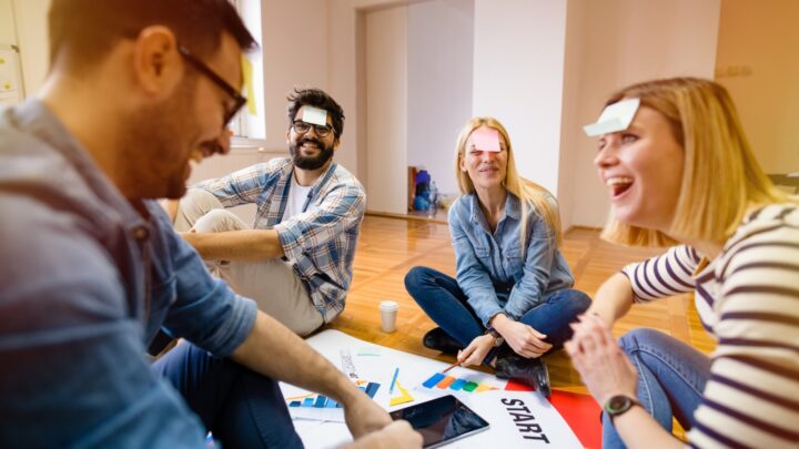 Four people sit on the floor laughing, each with a sticky note on their forehead. Papers, a tablet, and charts are spread in front of them, suggesting a playful group activity or team-building exercise in a bright room.