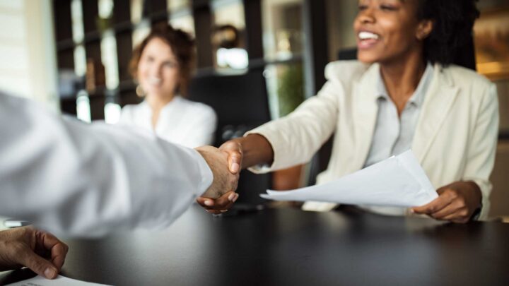 Two women sit at a table in an office, smiling as one of them shakes hands with a person across from her. One woman holds documents, and bookshelves are visible in the background.