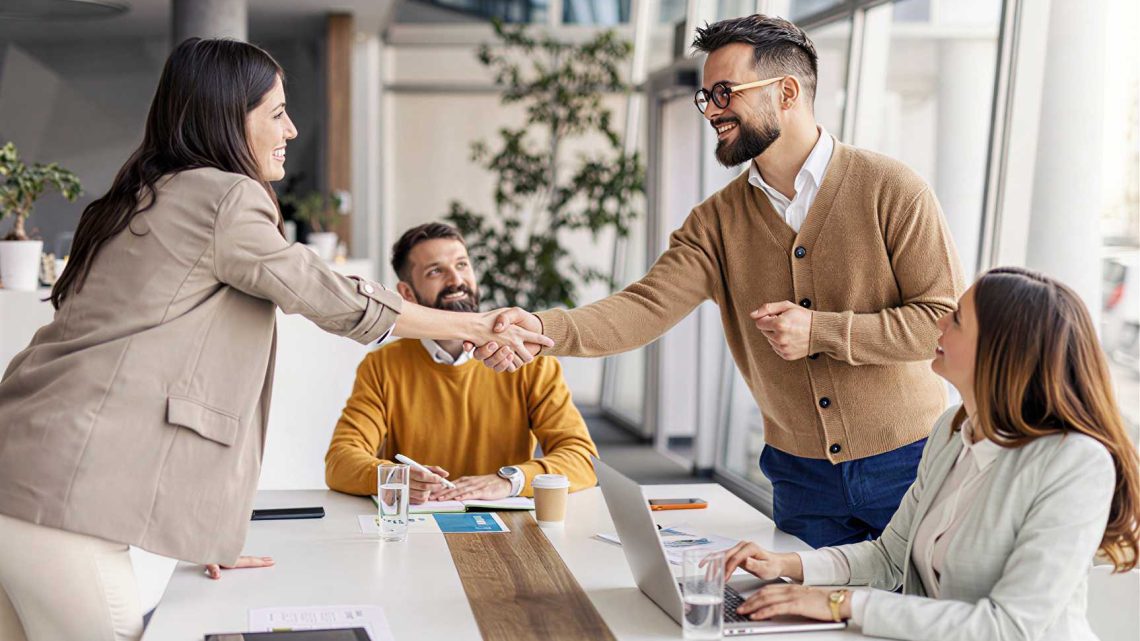 Four people in business attire sit around a meeting table. Two of them stand and shake hands, smiling. Laptops, papers, and glasses of water are on the table in a bright office space.