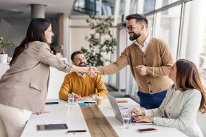 Four people in business attire sit around a meeting table. Two of them stand and shake hands, smiling. Laptops, papers, and glasses of water are on the table in a bright office space.