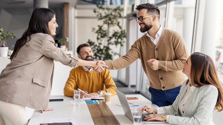 Four people in business attire sit around a meeting table. Two of them stand and shake hands, smiling. Laptops, papers, and glasses of water are on the table in a bright office space.
