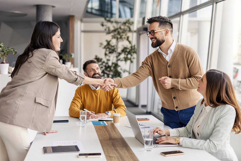 Four people in business attire sit around a meeting table. Two of them stand and shake hands, smiling. Laptops, papers, and glasses of water are on the table in a bright office space.